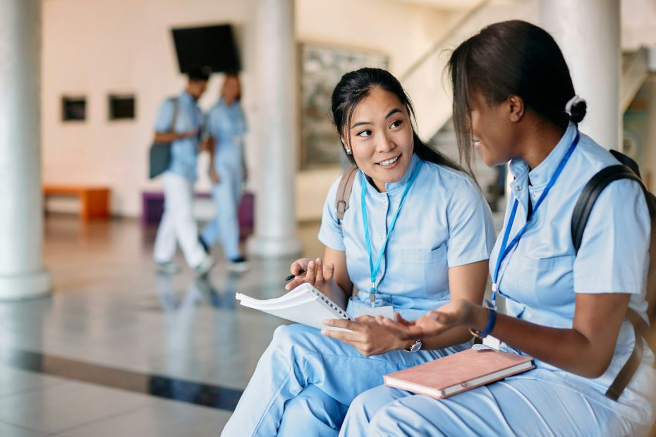 Online Nursing Programs - Asian nursing student and African-American friend talking while studying in hallway at medical university.
